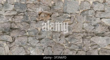 A detailed texture of a rough, weathered gray stone wall built with irregular blocks and mortar. Stock Photo