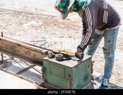 A construction worker wearing safety gloves and a sun hat uses a tape measure to mark steel rebar on a bending machin Stock Photo