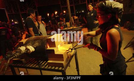 A woman working at a forge in a warmly lit blacksmith workshop at night, Burning Man, Nevada, USA Stock Photo