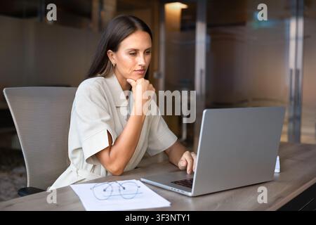 Businesswoman sitting at desk, reviewing documents, checking data on laptop Stock Photo