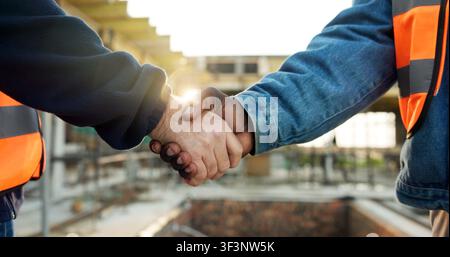 Shaking hands, men and construction workers on site for deal on building, maintenance or repairs. Scaffolding, industry and civil engineer with Stock Photo