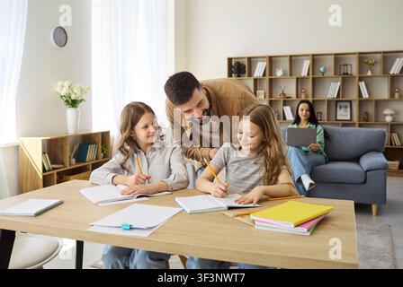 Father helping daughters with homework at home, mother working on laptop nearby Stock Photo
