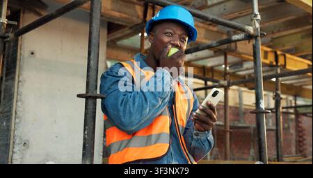 Apple, break and man at construction site with phone, fruit snack and PPE for building project. Black person, reflection and eating lunch outdoor with Stock Photo