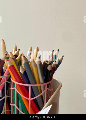 Bright colored pencils lean in a pink wire holder. Soft light highlights every tip and hue. Simple background emphasizes creativity and choice. Ideal Stock Photo