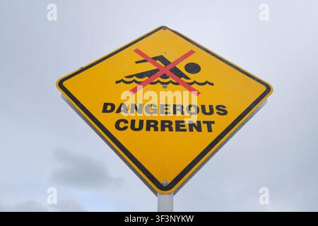 A yellow diamond warning sign that says, Dangerous Current. With swimmer graphic. On a cloudy day at Tamarama Beach near Sydney, Australia. Stock Photo