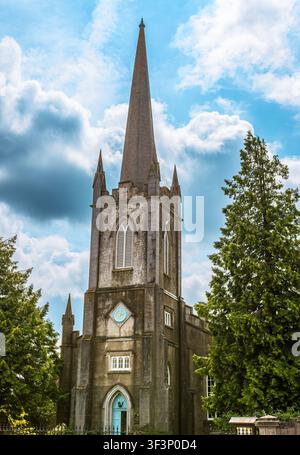Vertical image of a historic stone church in Ireland featuring a tall tower with a pointed spire, arched windows, a clock, and a blue entrance door, s Stock Photo