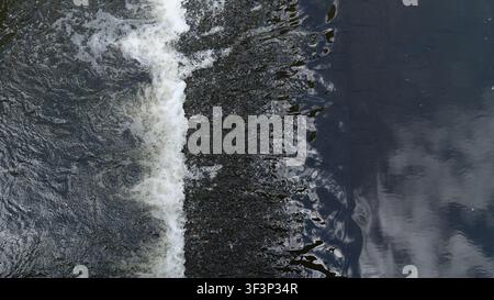 Top down view of flowing water streaming over a small dam or weir threshold creating white foam, splashes, and dark water reflections. Abstract aquati Stock Photo