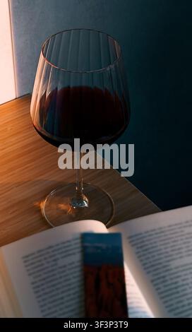 Glass of red wine placed on a wooden table beside an open book with a bookmark, soft lighting illuminating the scene from the side Stock Photo