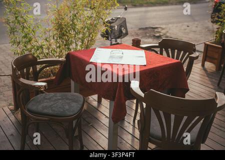 High-angle shot of an empty outdoor cafe table set with a red tablecloth, a white runner, an ashtray, and three wooden chairs. Selective focus. Stock Photo