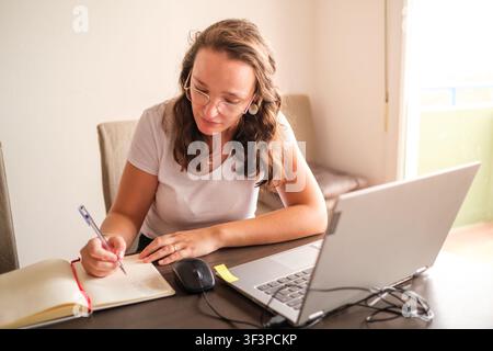 Woman writing in a notebook, studying or working remotely from home with a laptop Stock Photo