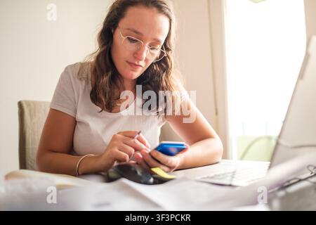Woman studying, checking smartphone, working remotely on laptop at house table Stock Photo