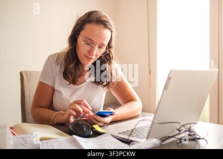 Woman focusing on her smartphone while using a laptop at a home office desk Stock Photo