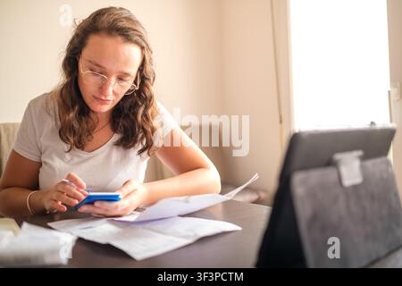 Woman calculating monthly expenses using a smartphone and paperwork at a home office desk Stock Photo