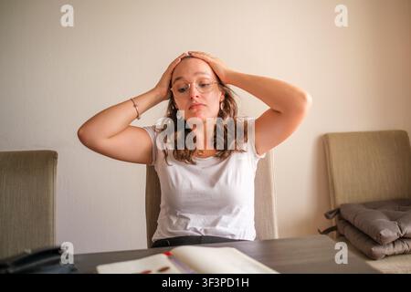Young woman sitting at a table feeling overwhelmed, stressed, and experiencing a headache Stock Photo