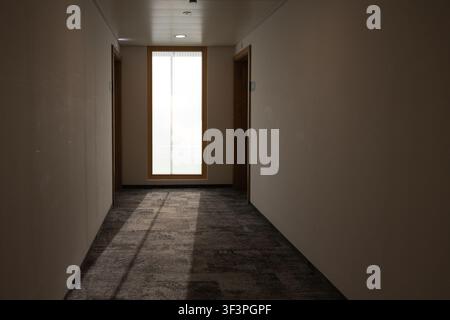 A dim, modern building hallway with grey carpeted flooring leading toward a tall window at the far end, casting natural sunlight across the floor Stock Photo