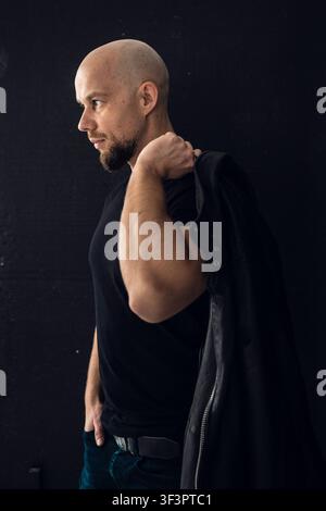 Man stands against dark background holding jacket over shoulder in casual pose Stock Photo