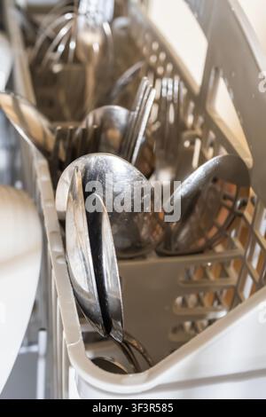A selective focus macro shot of clean metal spoons, forks, and a whisk inside a plastic cutlery basket of an open dishwasher. Stock Photo