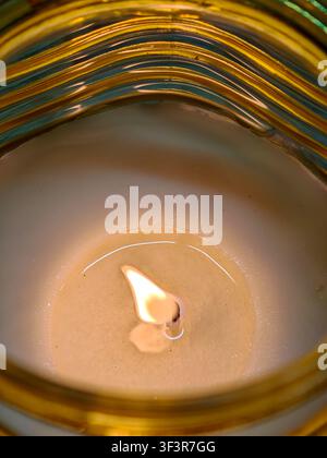 Warm light from a burning candle creates a serene atmosphere, reflecting on the melting wax surface and creating abstract patterns on the glass holder Stock Photo