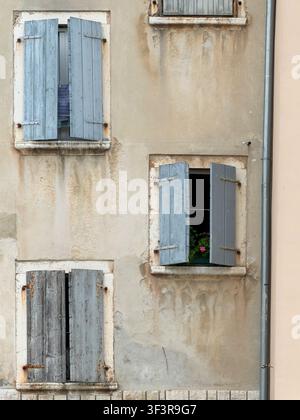 Faded blue-grey wooden shutters frame windows on a weathered stucco building, with potted pink flowers brightening one open windowsill. Stock Photo