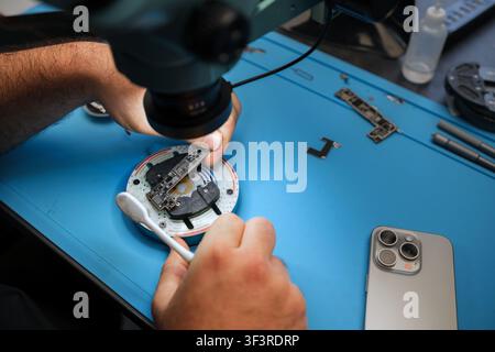 Technician repairs electronic device under microscope with tools and smartphone on blue work surface in a modern workshop setting Stock Photo