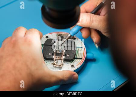 Technician repairs smartphone circuit board under magnifying lens on a blue work surface with precision tools and equipment visible Stock Photo