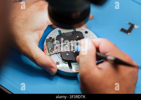 Technician repairs electronic circuit board under magnifying lens with precision tools on blue work surface in a well-lit environment Stock Photo
