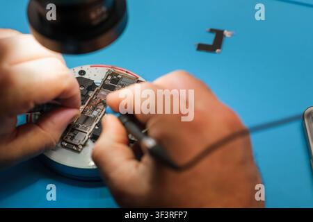 Technician repairs smartphone circuit board under magnifying lamp on blue work surface with tools and components visible in the background Stock Photo
