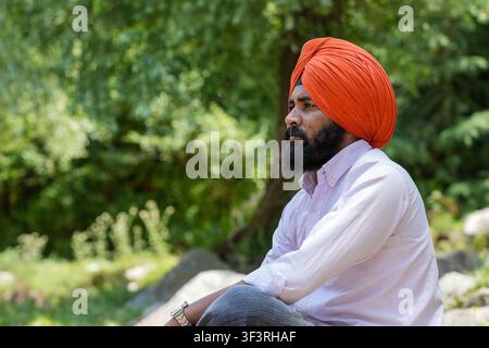 Closeup side view of a young Sikh man wearing an orange turban, looking away thoughtfully against a blurred background of lush green trees in Himachal. Stock Photo