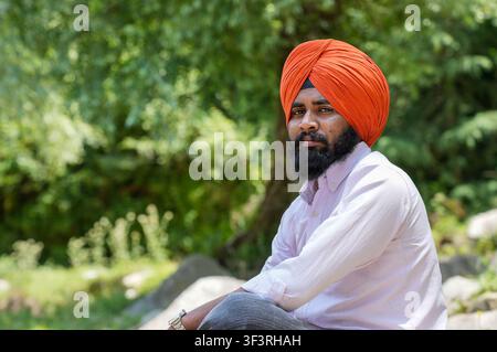 Closeup portrait of a young Sikh man wearing an orange turban, looking directly at the camera while sitting against a blurred background of lush green. Stock Photo