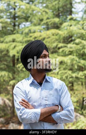 Vertical portrait of a young Sikh man wearing a black turban and a blue shirt, standing with arms crossed while looking away. Stock Photo