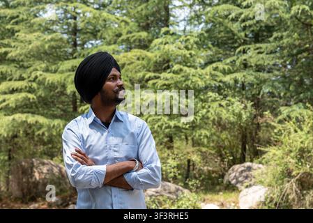 A happy young Sikh man wearing a black turban and light blue shirt, stands in a lush forest setting with his arms crossed, looking toward the side. Stock Photo