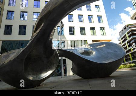 A large abstract metal sculpture stands in front of modern office buildings in a city setting. Bright daylight and a wide-angle perspective emphasize Stock Photo