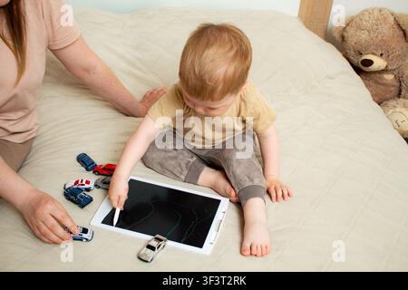 Toddler drawing on tablet using stylus, mom guiding his hand on tablet screen. Early creative development through shared activity. Parent child game s Stock Photo