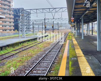 A deserted railway station platform in Japan on an overcast, rainy day. The image shows multiple parallel train tracks extending into the distance Stock Photo