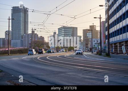 Urban street scene in Japan featuring curved tram tracks running along a multi-lane road with several vehicles including white vans and passenger Stock Photo
