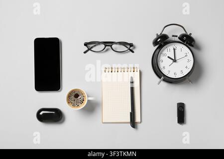 Flat lay of office essentials including a smartphone, coffee, notebook, alarm clock, and glasses on a light gray background Stock Photo