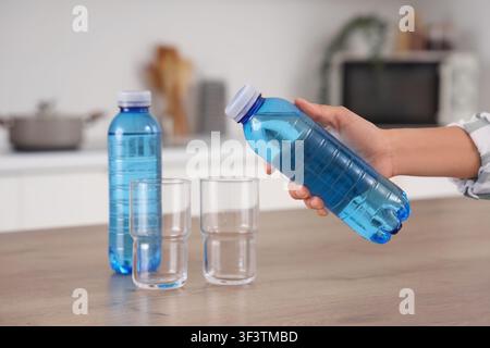 Person holding a blue plastic water bottle with two empty glasses and Another bottle on a kitchen counter, Another full blue water bottle stands nearb Stock Photo