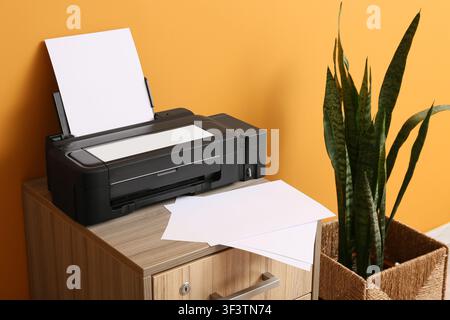 Black inkjet printer with blank white paper on a wooden cabinet next to a snake plant in a woven basket, Additional sheets of paper are stacked on the Stock Photo