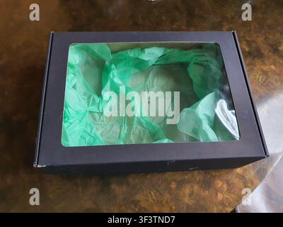 A black rectangular cardboard gift box with a clear plastic window in the lid, revealing crumpled green tissue paper inside. The box sits on a brown Stock Photo