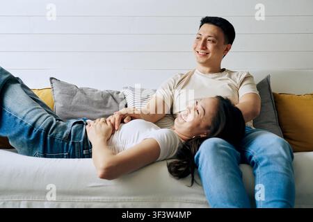 Smiling man and woman relaxing together on a cozy sofa, enjoying a calm weekend at home with soft pillows and warm light, expressing comfort, intimacy Stock Photo