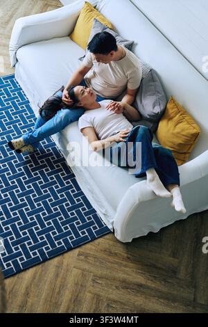 Overhead view of a couple relaxing together on a white sofa at home, sharing a quiet moment. Man rests beside woman as they hold hands in a cozy livin Stock Photo