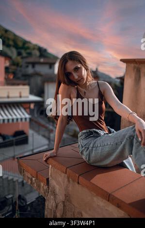 Young woman posing for a street-style portrait on an Italian rooftop terrace at sunset, overlooking colorful houses and hills, with soft evening light Stock Photo