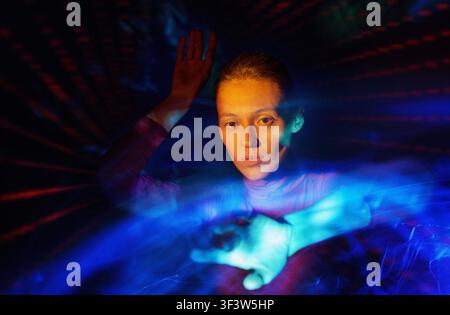 Futuristic portrait of a young woman surrounded by vivid blue and red light trails, reaching forward in a dark studio, evoking virtual reality, immers Stock Photo
