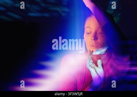 Young woman in vivid neon blue and pink light interacts with a futuristic virtual reality interface, reaching and touching glowing holograms in a dark Stock Photo