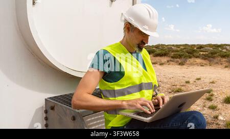 Male renewable energy technician and engineer in hard hat and reflective vest uses a laptop to inspect and document wind turbine maintenance in a dese Stock Photo