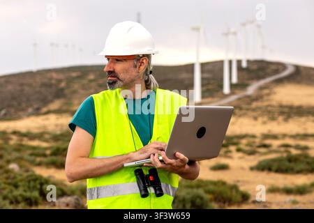 Male renewable energy engineer in hard hat and reflective vest uses a laptop to inspect wind turbines at a wind farm, monitoring performance and maint Stock Photo