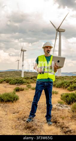 Male renewable energy technician and engineer in safety vest and hard hat checks wind turbine performance on site using laptop and binoculars under cl Stock Photo