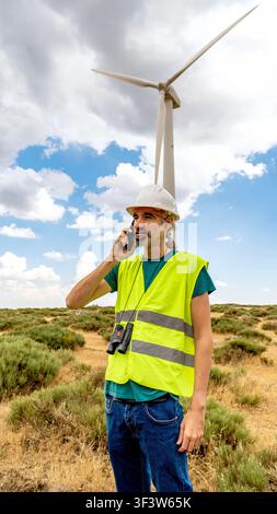 Renewable energy technician and engineer wearing hard hat and safety vest talks on smartphone at a wind farm, inspecting turbine performance in a rura Stock Photo