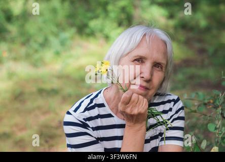 Close-up portrait of a thoughtful senior woman with short gray hair holding a yellow wildflower, sitting outdoors in a peaceful green forest with soft Stock Photo
