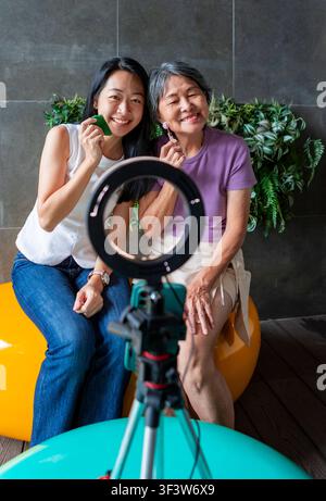 Smiling mother and adult daughter sit together in a modern home studio, holding facial massage tools while recording a skincare tutorial with a ring l Stock Photo